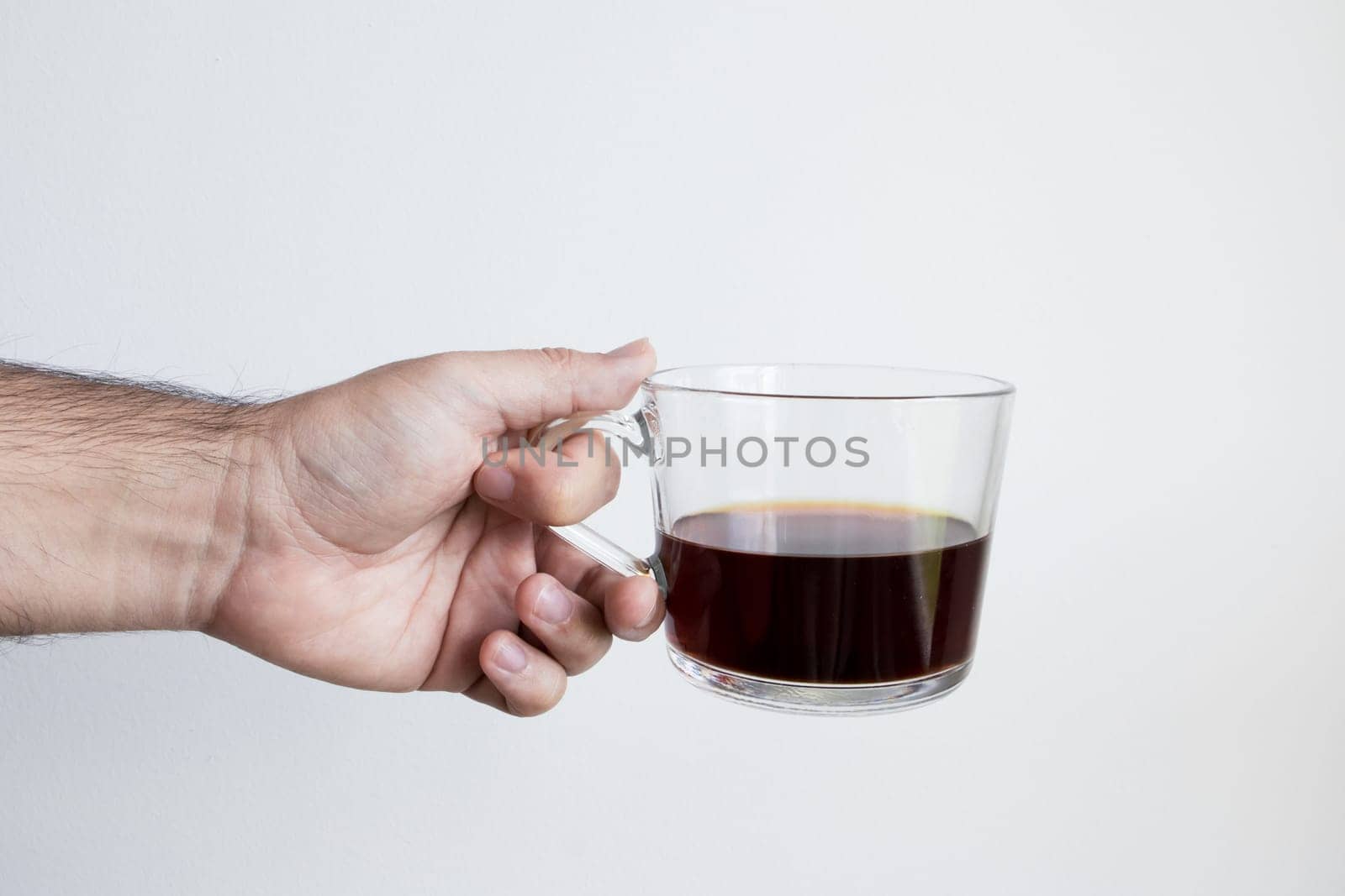 Caucasian male hand holding clear coffee cup against plain background.