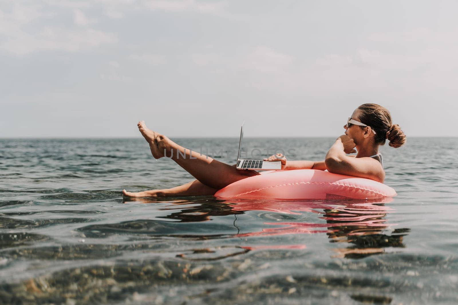 A woman is floating in a pink inflatable raft with a laptop in her hand. She is smiling and she is enjoying her time in the water