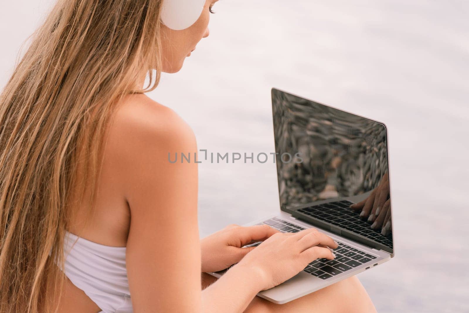 A woman is sitting on a beach with a laptop in her lap. She is wearing headphones and she is listening to music
