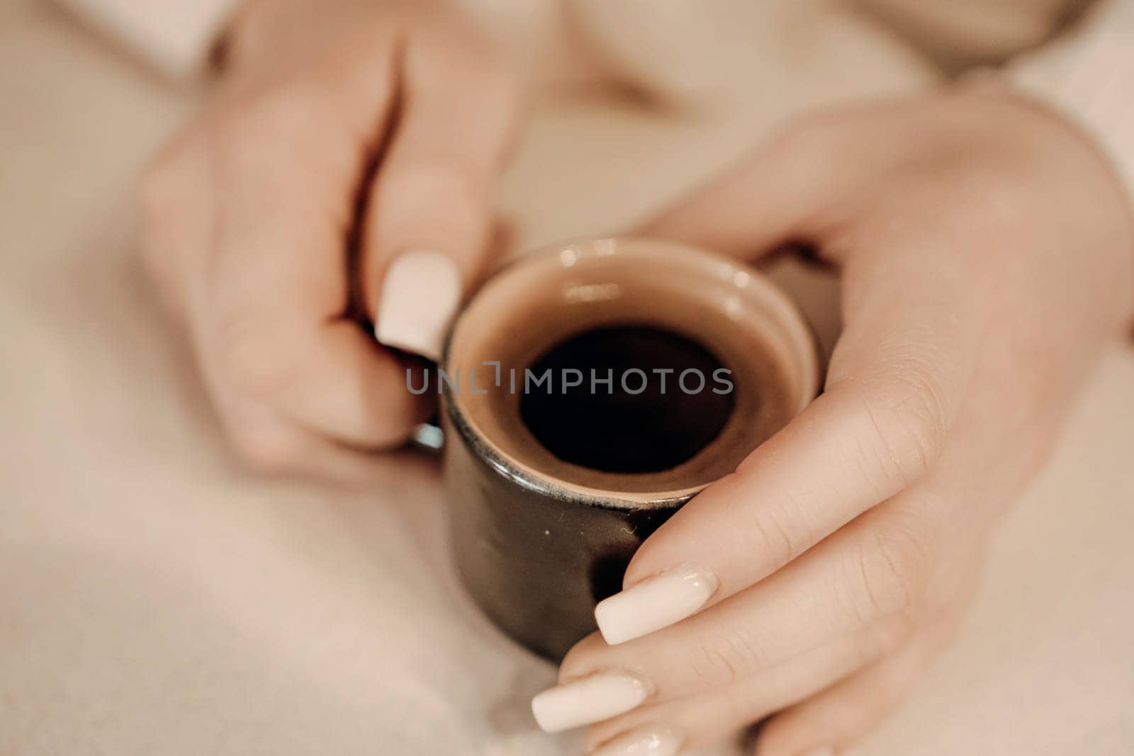 Close-up of two hands gently cradling a small, dark-colored cup filled with dark coffee. Cream-colored nails are visible.