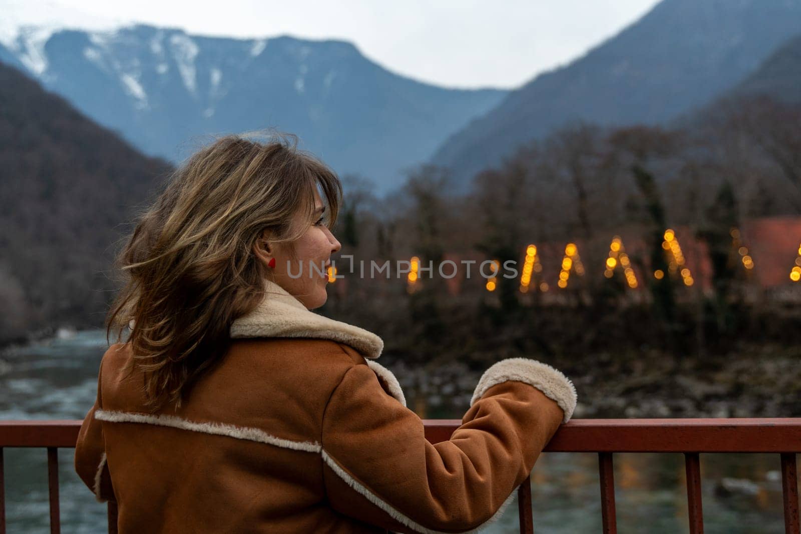 Woman in brown shearling jacket stands on bridge, gazing at illuminated building nestled in mountainous valley. River flows below.