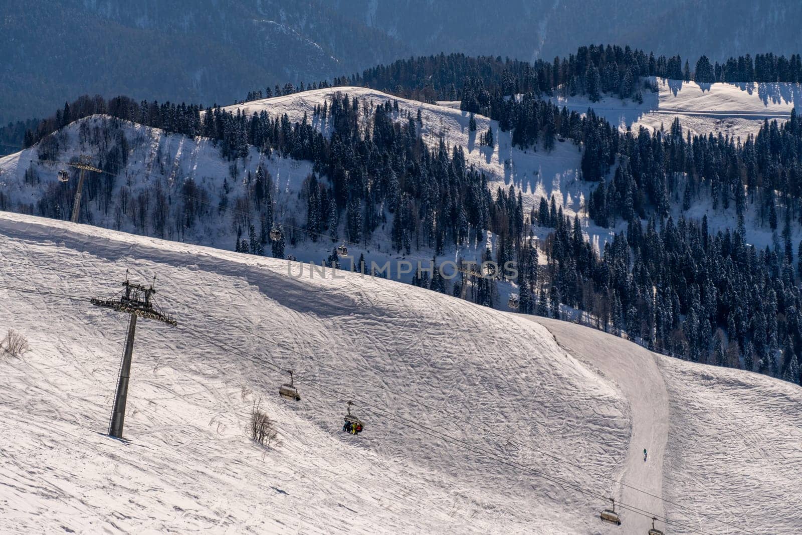Snow-covered mountain slope with a ski lift and sparsely populated ski runs. Trees line the hillsides under a clear sky. Winter landscape.
