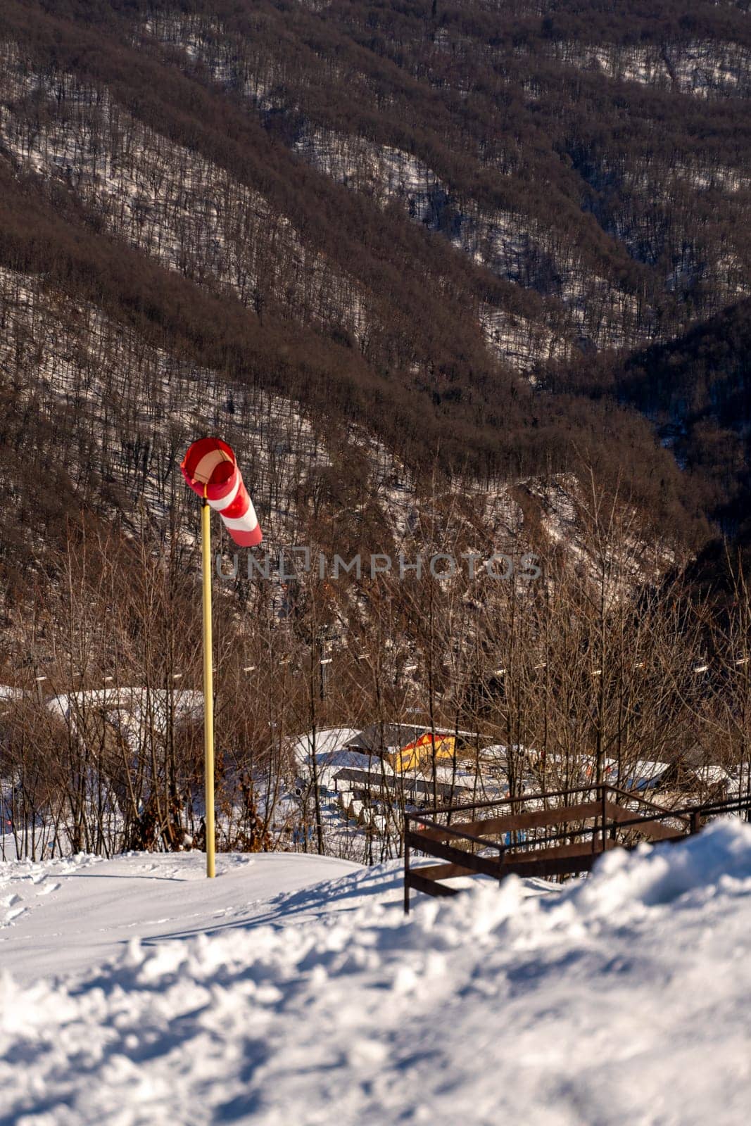 Red and white windsock indicates wind direction on a snowy mountainside. A small building and wooden fence are visible in the background. Winter landscape.