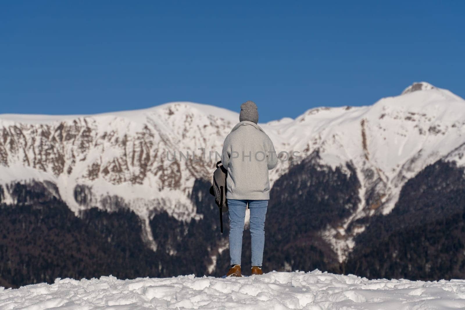 Person in light gray jacket and jeans stands on snowy mountain peak, gazing at snow-covered mountain range under a clear blue sky. Back view.