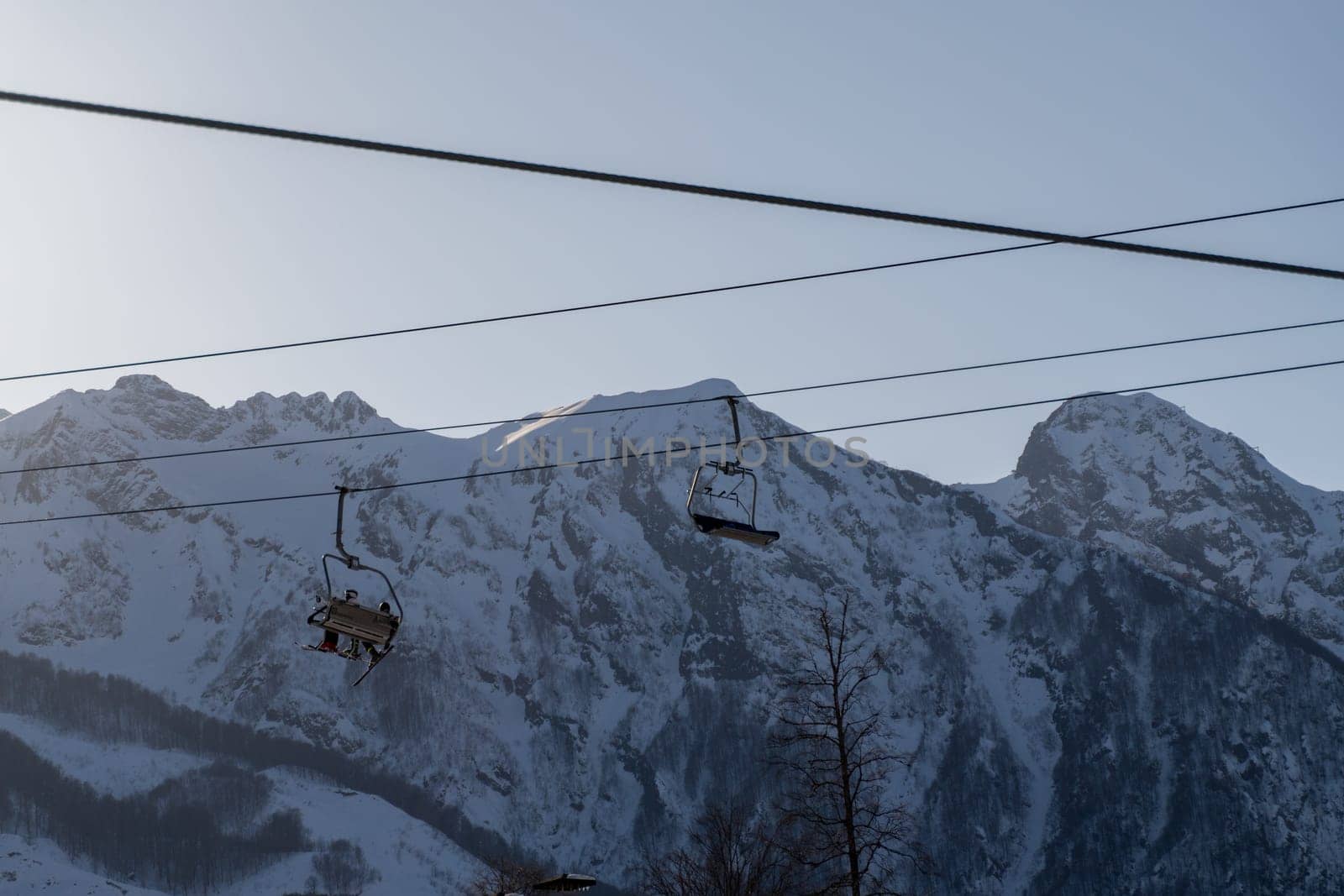 Two empty ski lift chairs hang against a backdrop of snow-covered mountains under a pale sky. Wires stretch across the scene.
