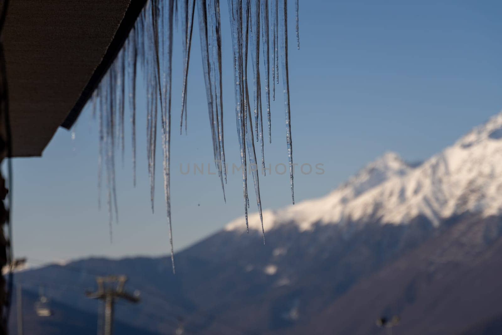 Numerous icicles hang from a roof's edge, a stark contrast to the snow-capped mountains in the background under a clear blue sky.