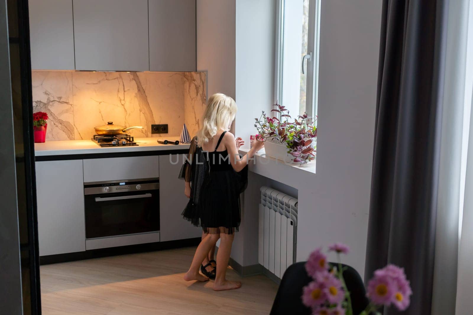 Blonde woman in black dress stands barefoot in modern kitchen, near window with flowers, looking outside. Countertop shows food prep.