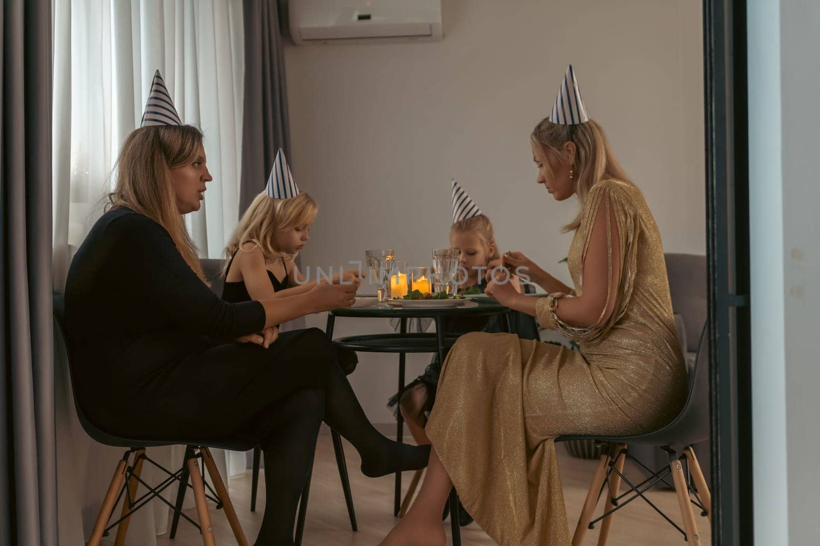 Three women and a baby girl celebrate a birthday. They wear party hats and sit around a small table with candles, seemingly enjoying a meal or small gifts.