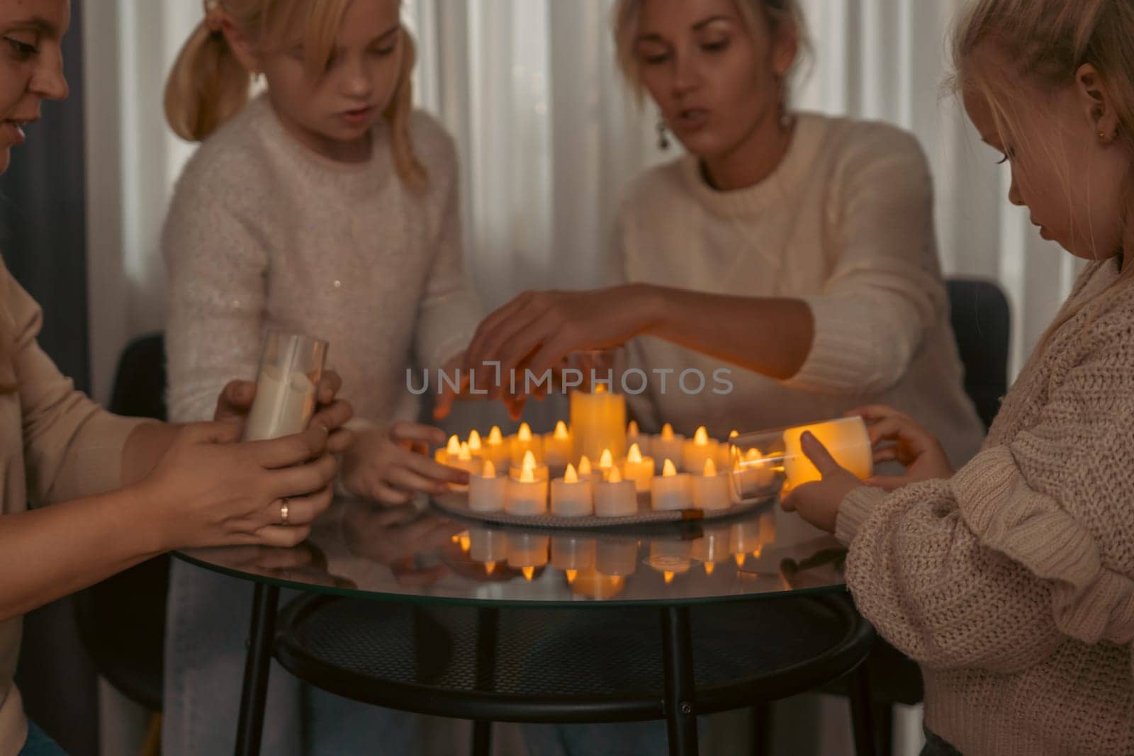 A woman and three girls light numerous small, flickering candles arranged on a circular glass table. Warm light illuminates their faces and hands as they work together.