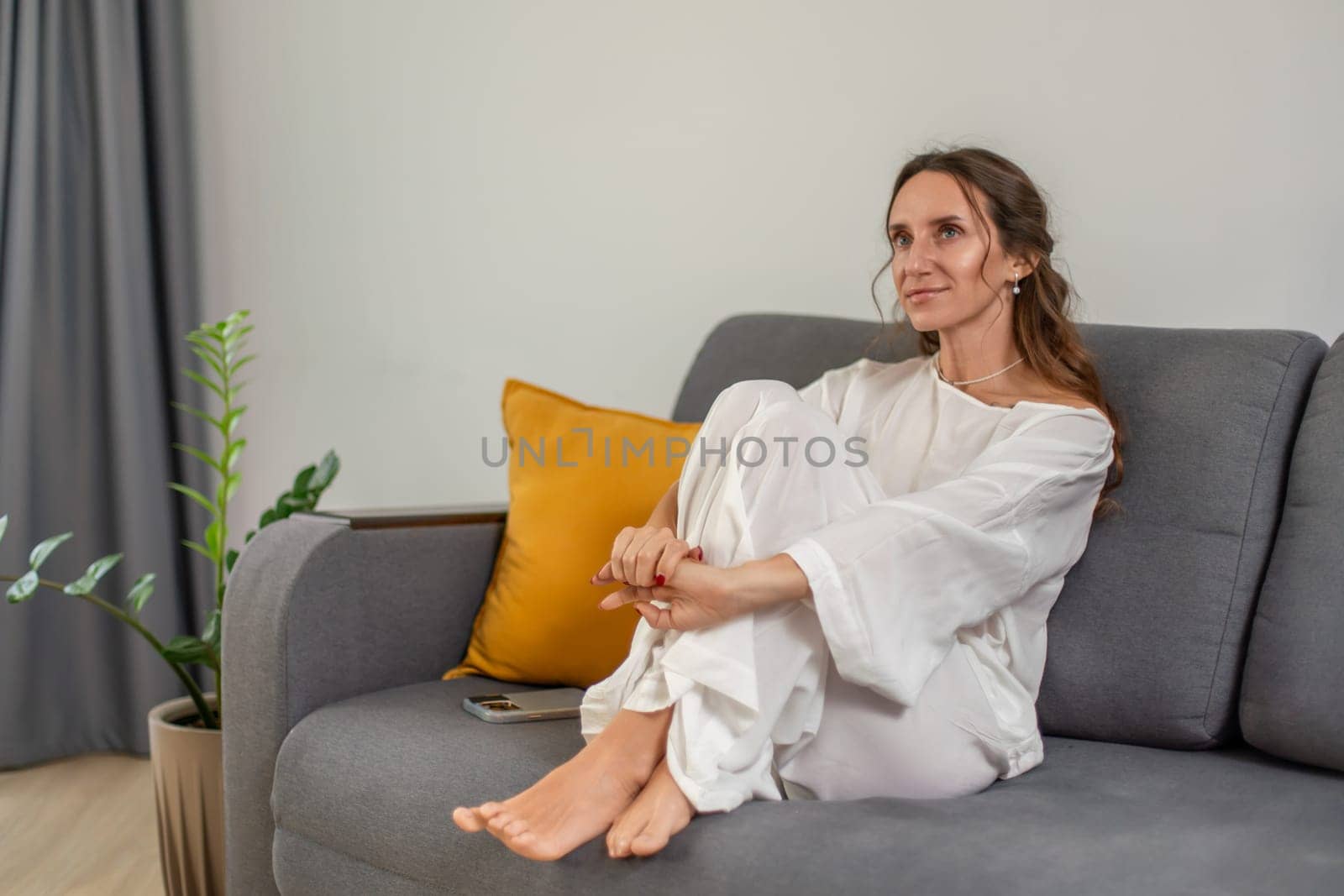 Woman in white loose clothing sits barefoot on a grey couch, legs crossed, hands clasped. A mustard yellow pillow rests beside her. Serene expression.