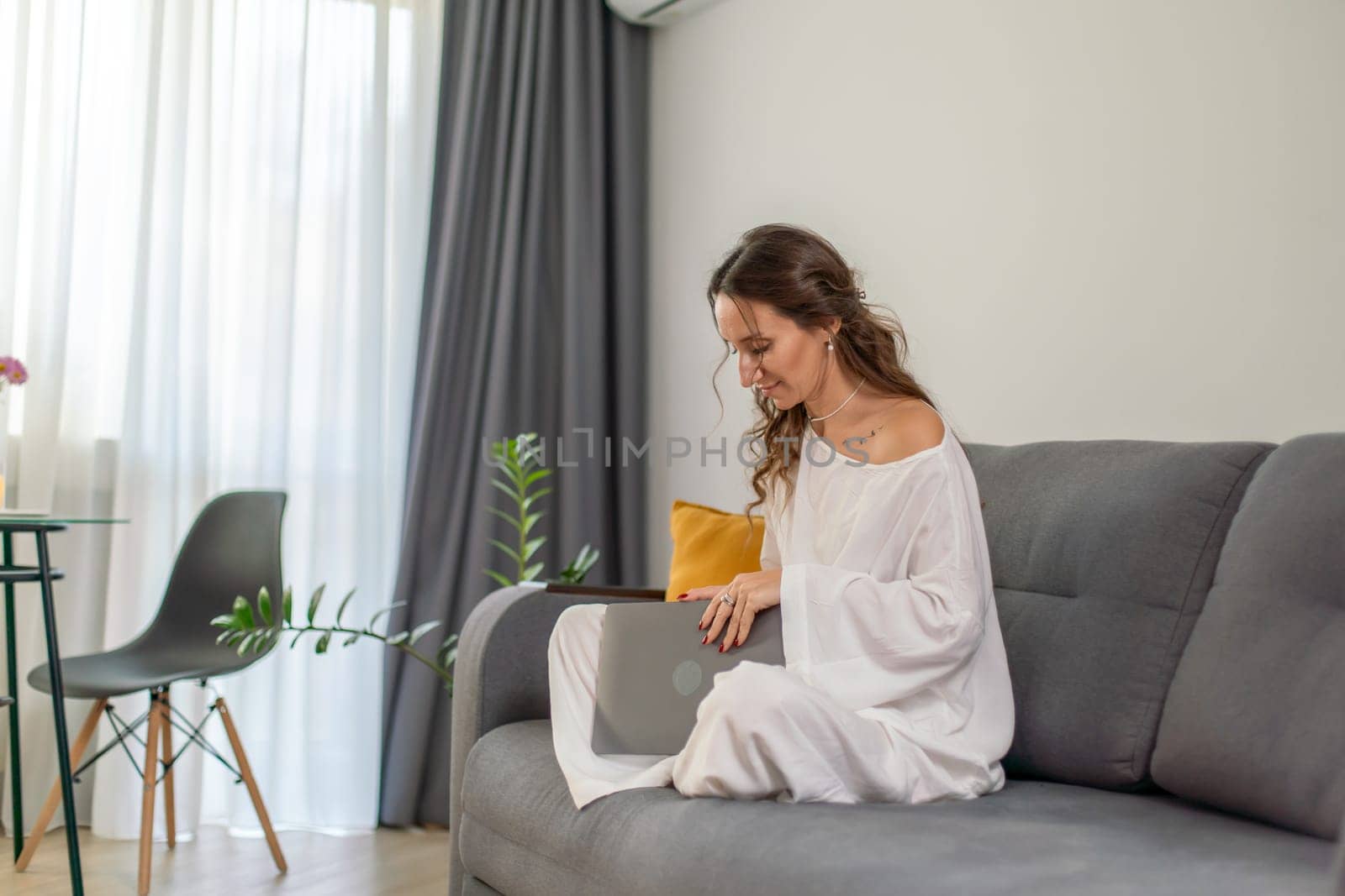 Woman in white pajamas sits on a gray couch in a modern apartment, examining a yellow object. Natural light streams through sheer curtains.