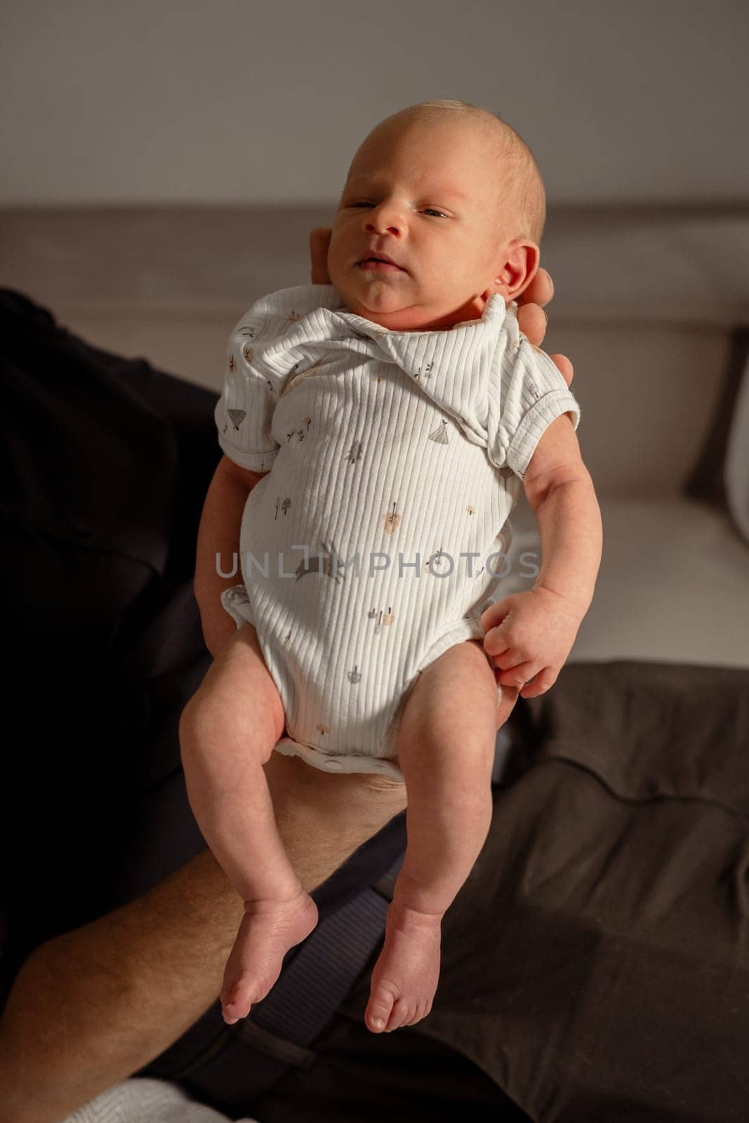 A small baby in a cozy indoor setting, gently held in a hand, wears a soft onesie and has a calm, tender expression.