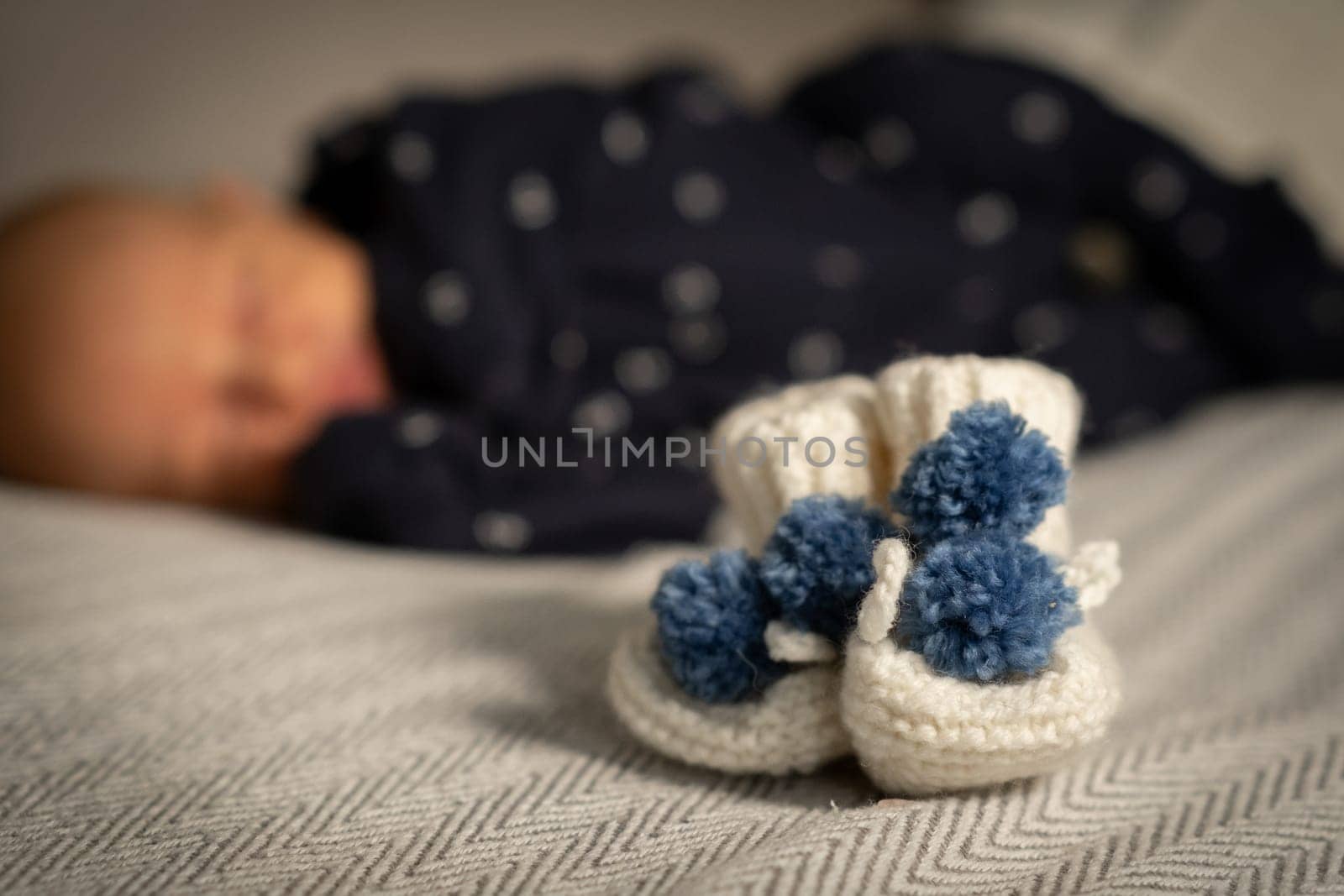 Soft white booties with blue pom poms rest on a blanket while a sleeping baby lies peacefully in the background of a serene indoor space
