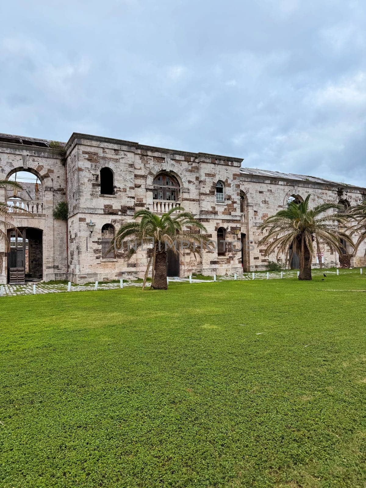Weathered stone ruins of a historic colonial-era building in Bermuda, surrounded by green lawn and palm trees under cloudy sky.