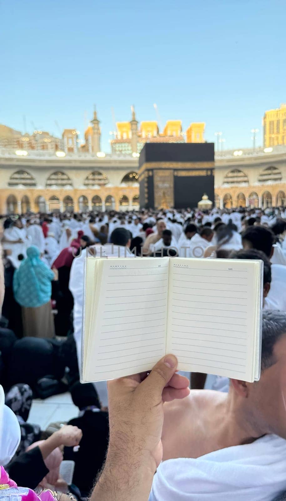 A pilgrim holds a small notebook with a heart doodle in front of the Kaaba in Mecca, Saudi Arabia. The sacred site is surrounded by worshippers performing Hajj or Umrah under a clear blue sky.