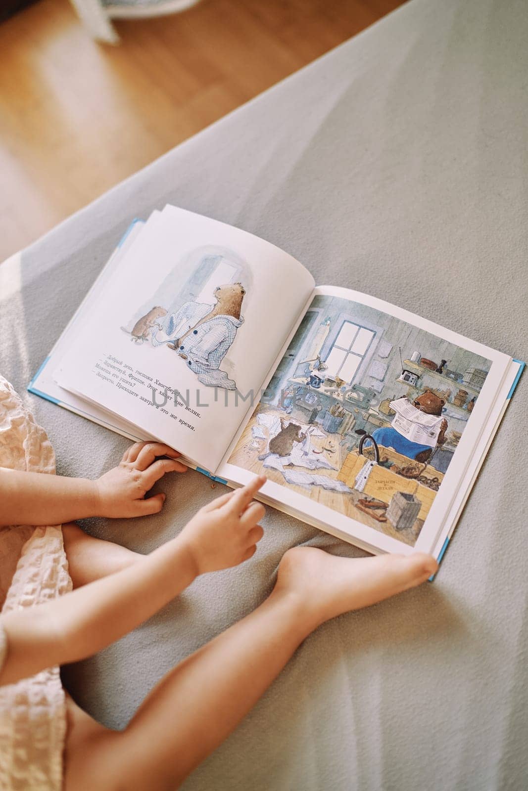 Little girl sits on her bed with a book about beavers and runs her finger over the pictures. Cropped. Faceless. High quality photo