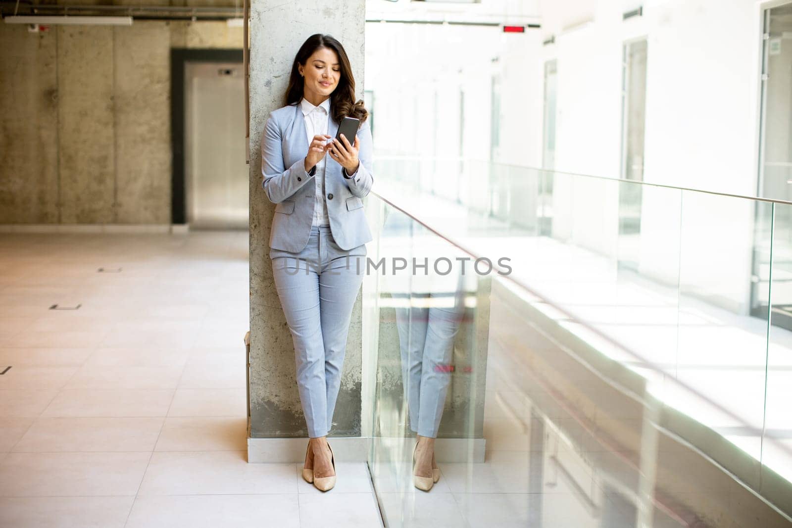 Pretty young business woman using mobile phone in the office hallway
