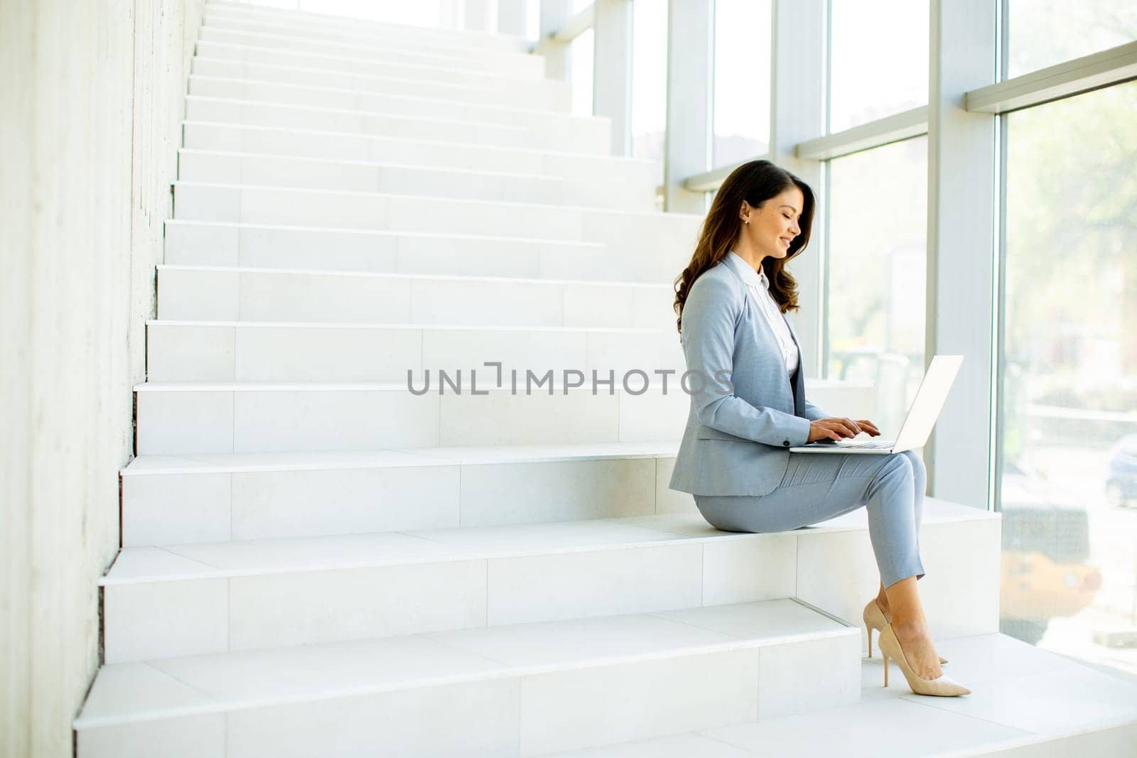 Pretty young business woman sitting on the stairs on office hallway and working on laptop