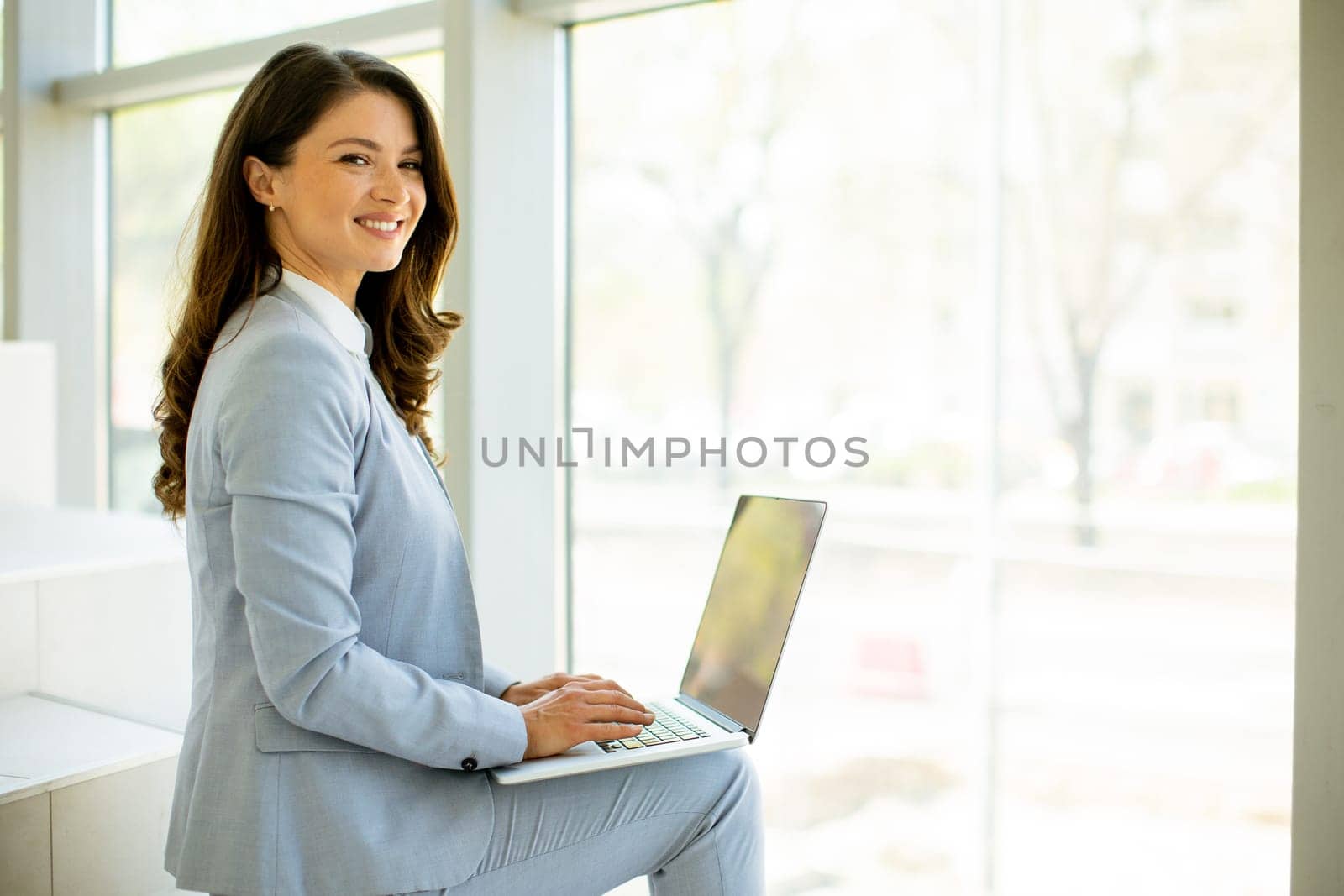 Pretty young business woman sitting on the stairs on office hallway and working on laptop