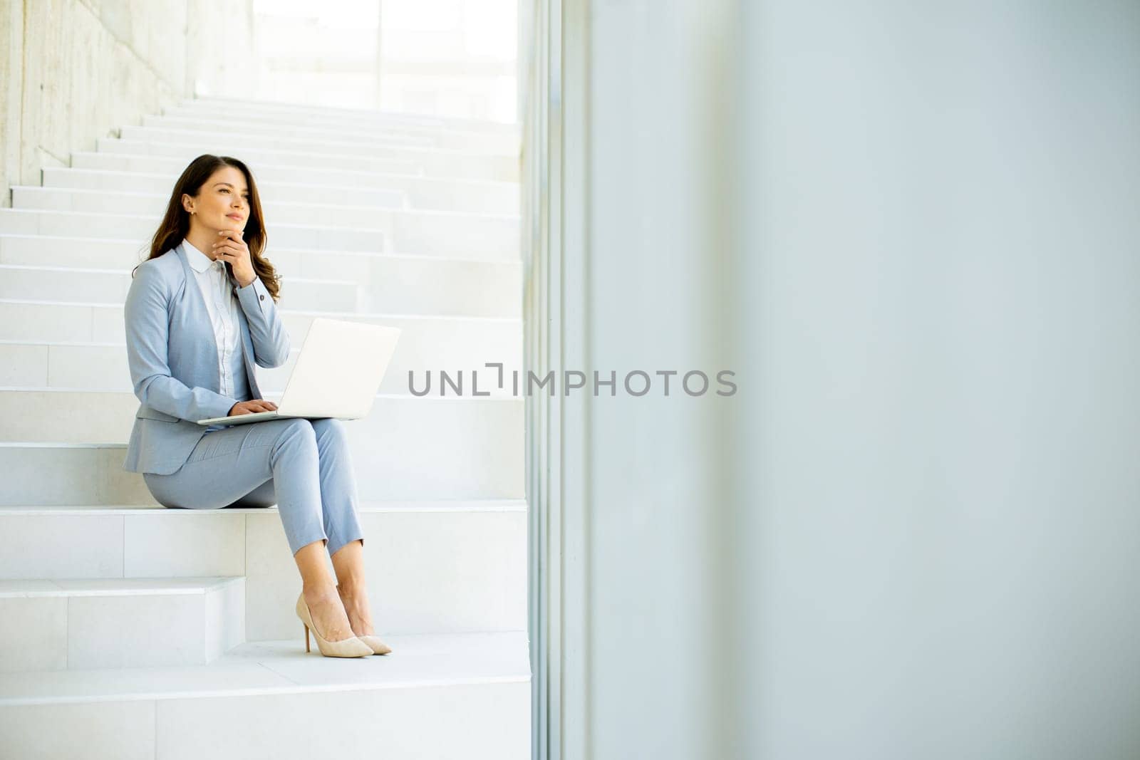 Pretty young business woman sitting on the stairs on office hallway and working on laptop
