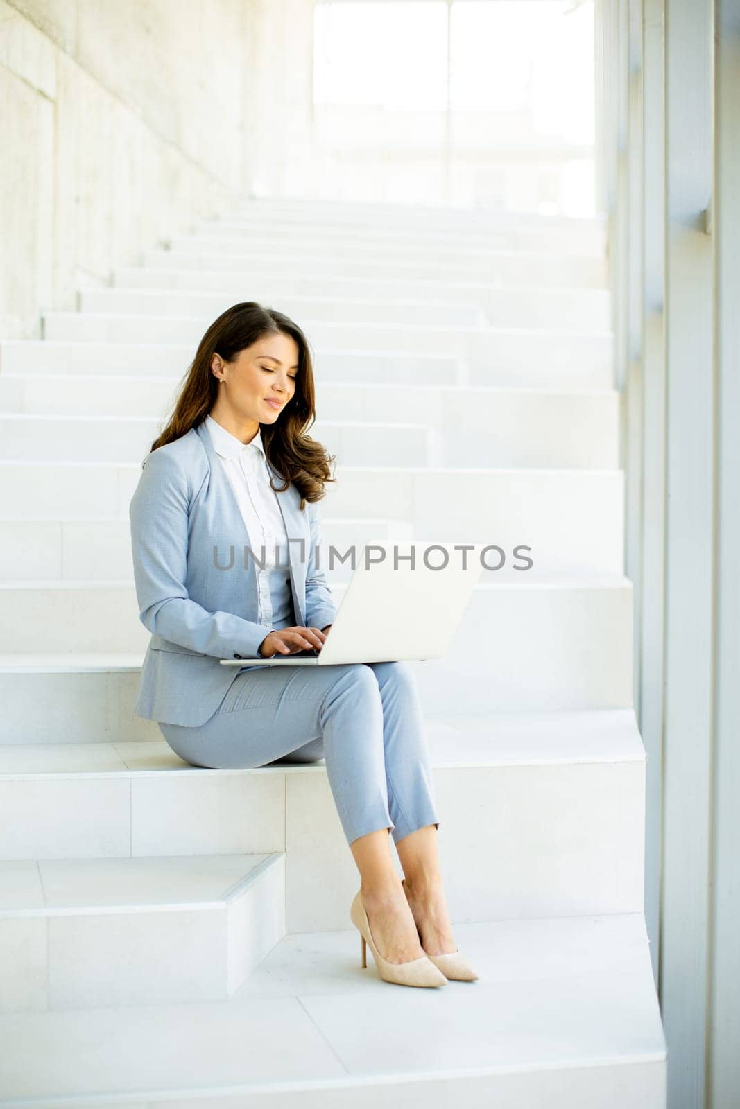 Pretty young business woman sitting on the stairs on office hallway and working on laptop
