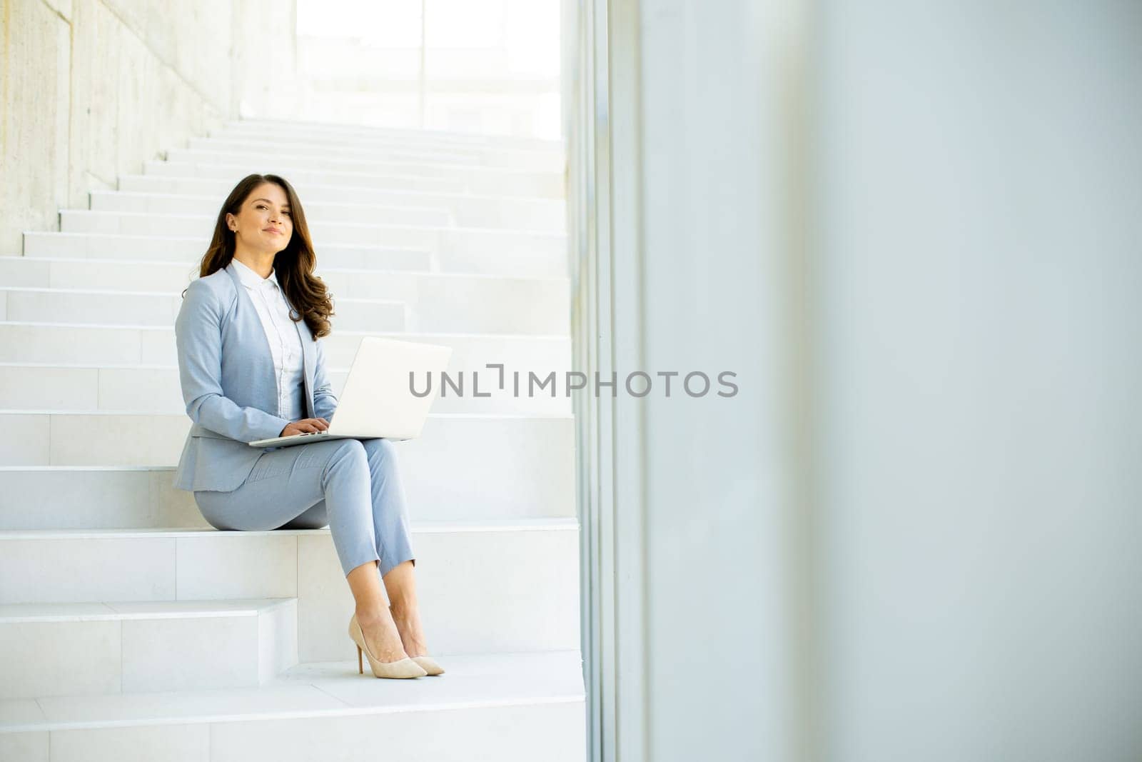 Pretty young business woman sitting on the stairs on office hallway and working on laptop