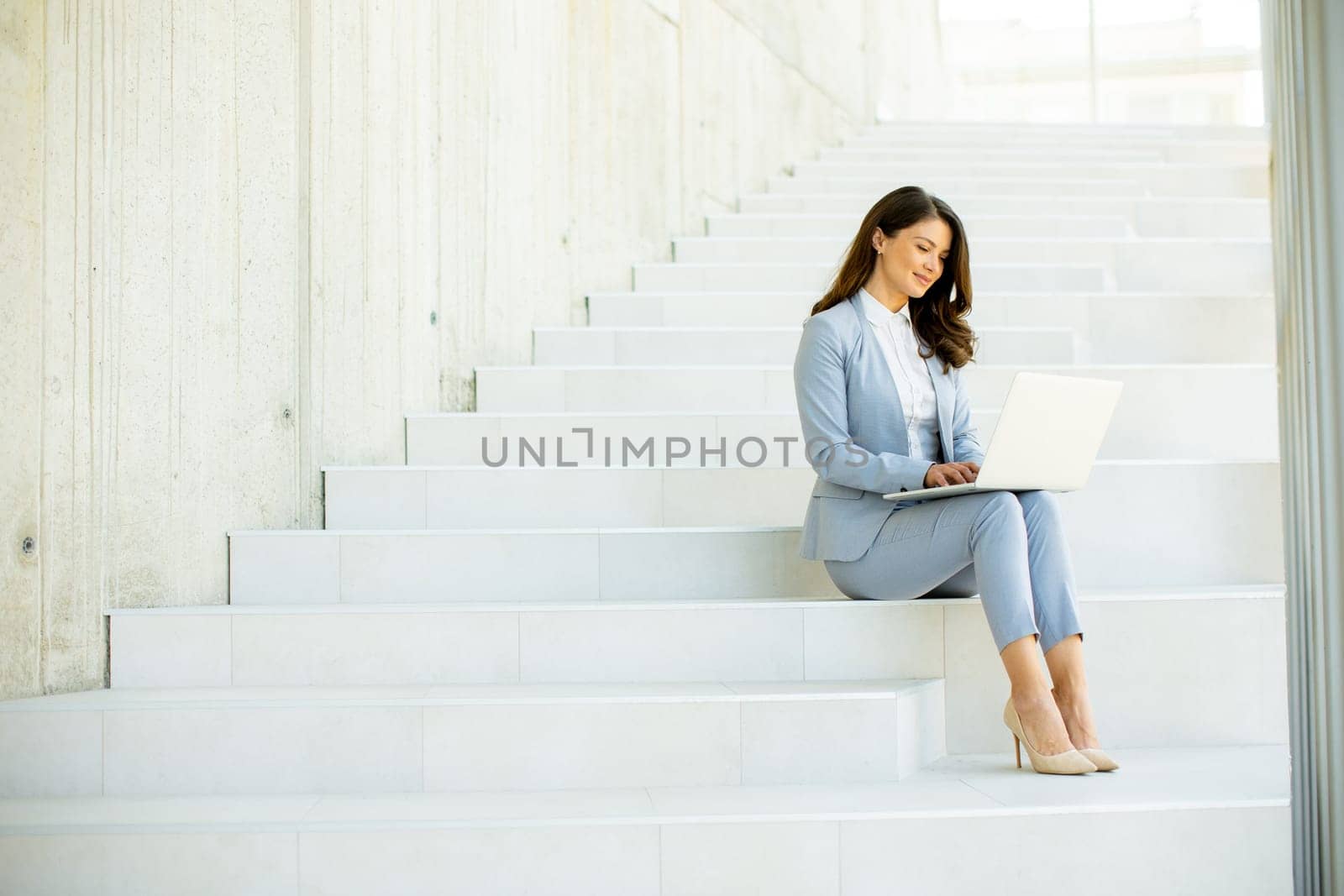 Pretty young business woman sitting on the stairs on office hallway and working on laptop