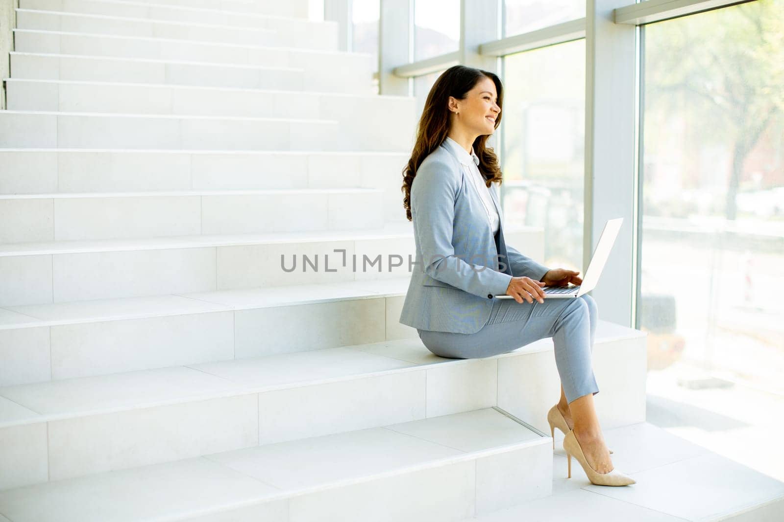 Pretty young business woman sitting on the stairs on office hallway and working on laptop