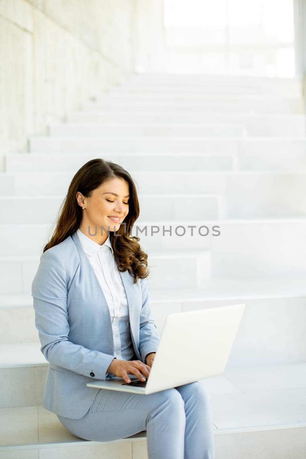 Pretty young business woman sitting on the stairs on office hallway and working on laptop