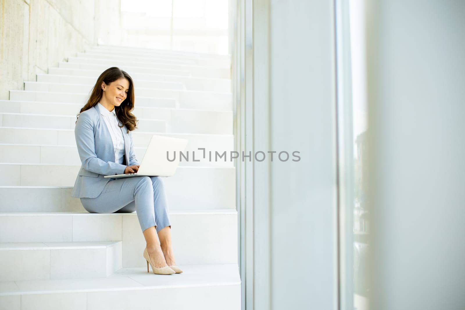 Pretty young business woman sitting on the stairs on office hallway and working on laptop