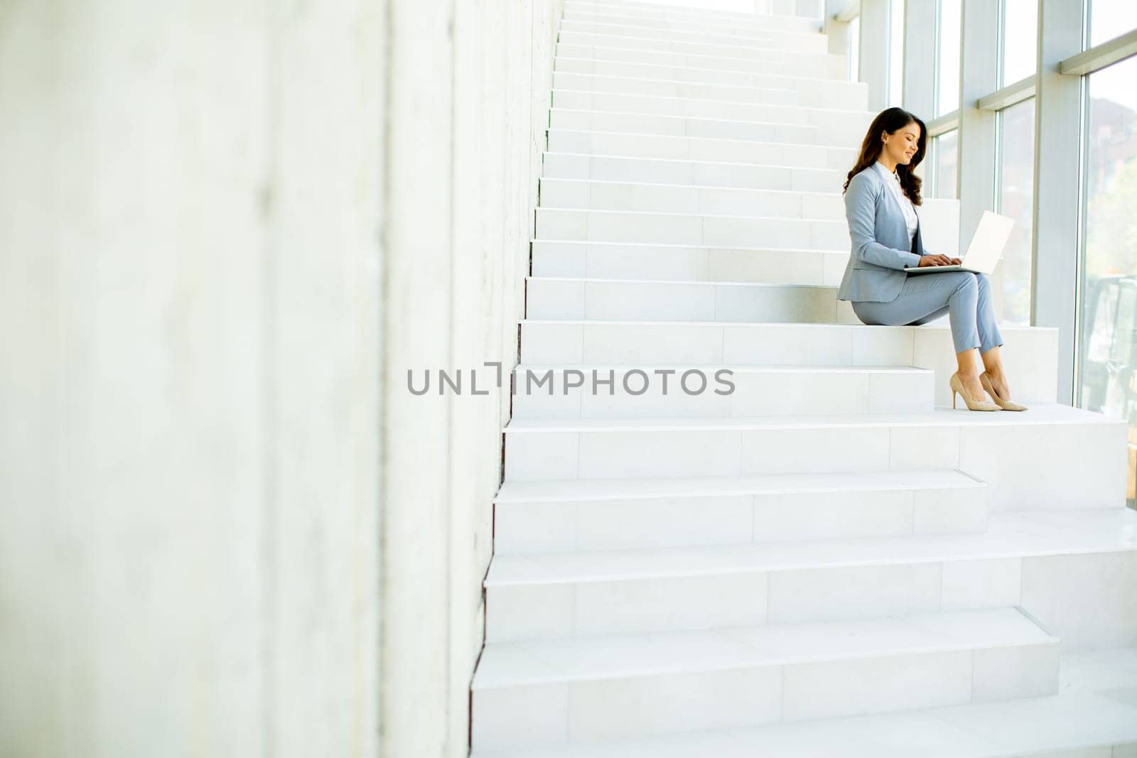 Pretty young business woman sitting on the stairs on office hallway and working on laptop