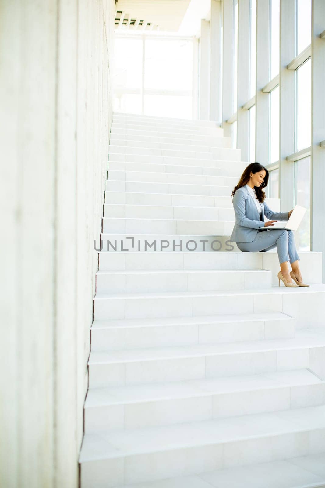 Pretty young business woman sitting on the stairs on office hallway and working on laptop