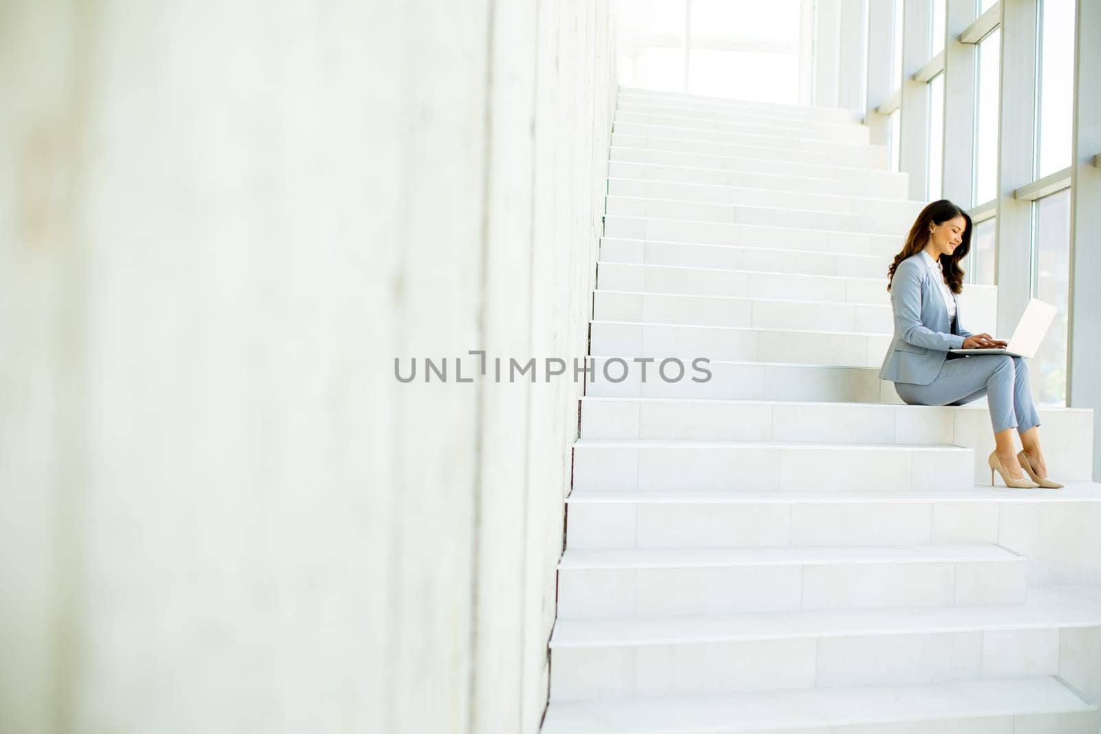Pretty young business woman sitting on the stairs on office hallway and working on laptop