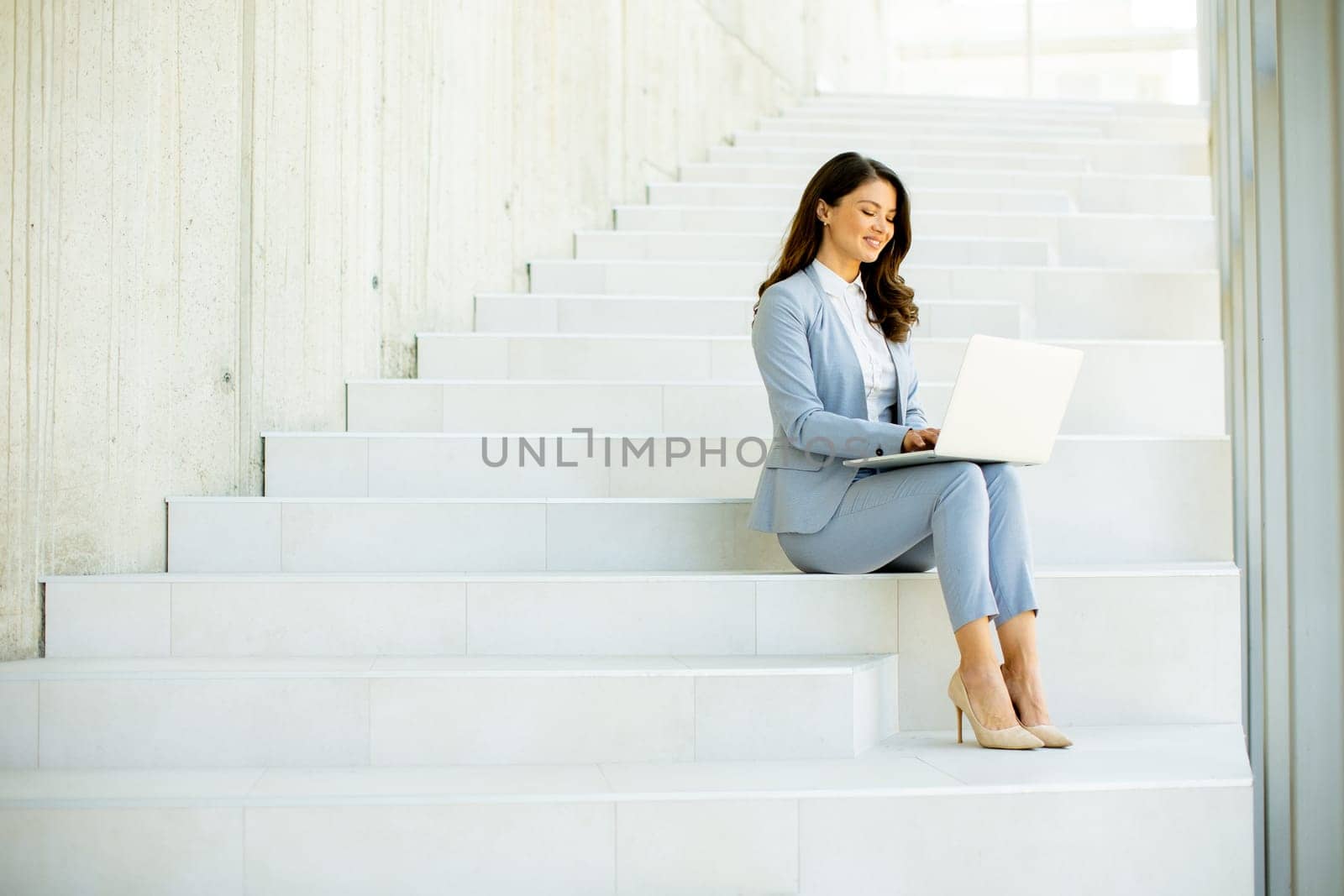Pretty young business woman sitting on the stairs on office hallway and working on laptop