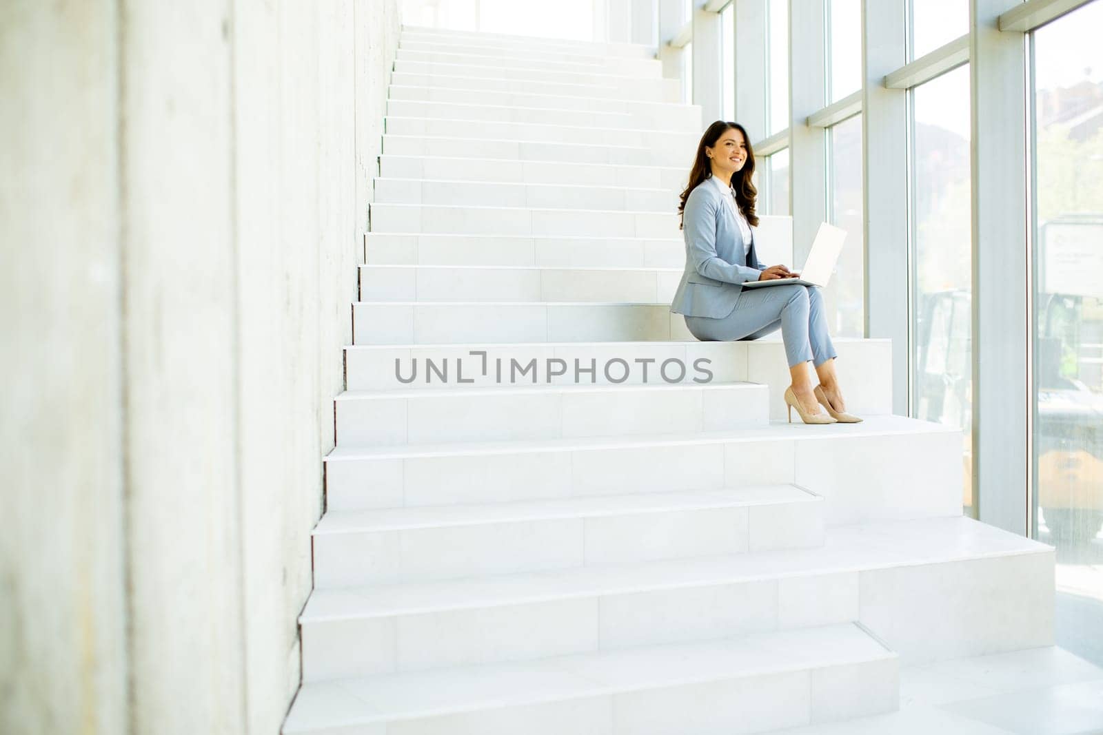 Pretty young business woman sitting on the stairs on office hallway and working on laptop