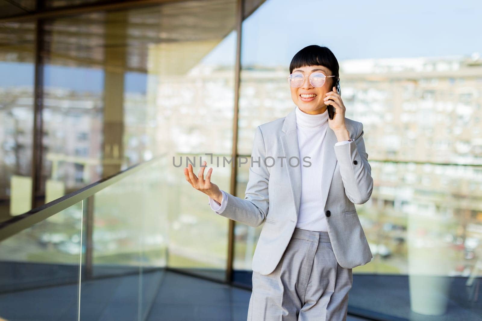 A professional Asian woman in a stylish suit chats joyfully on her phone within a sleek office building with large windows. Her bright smile reflects her positive energy and success.