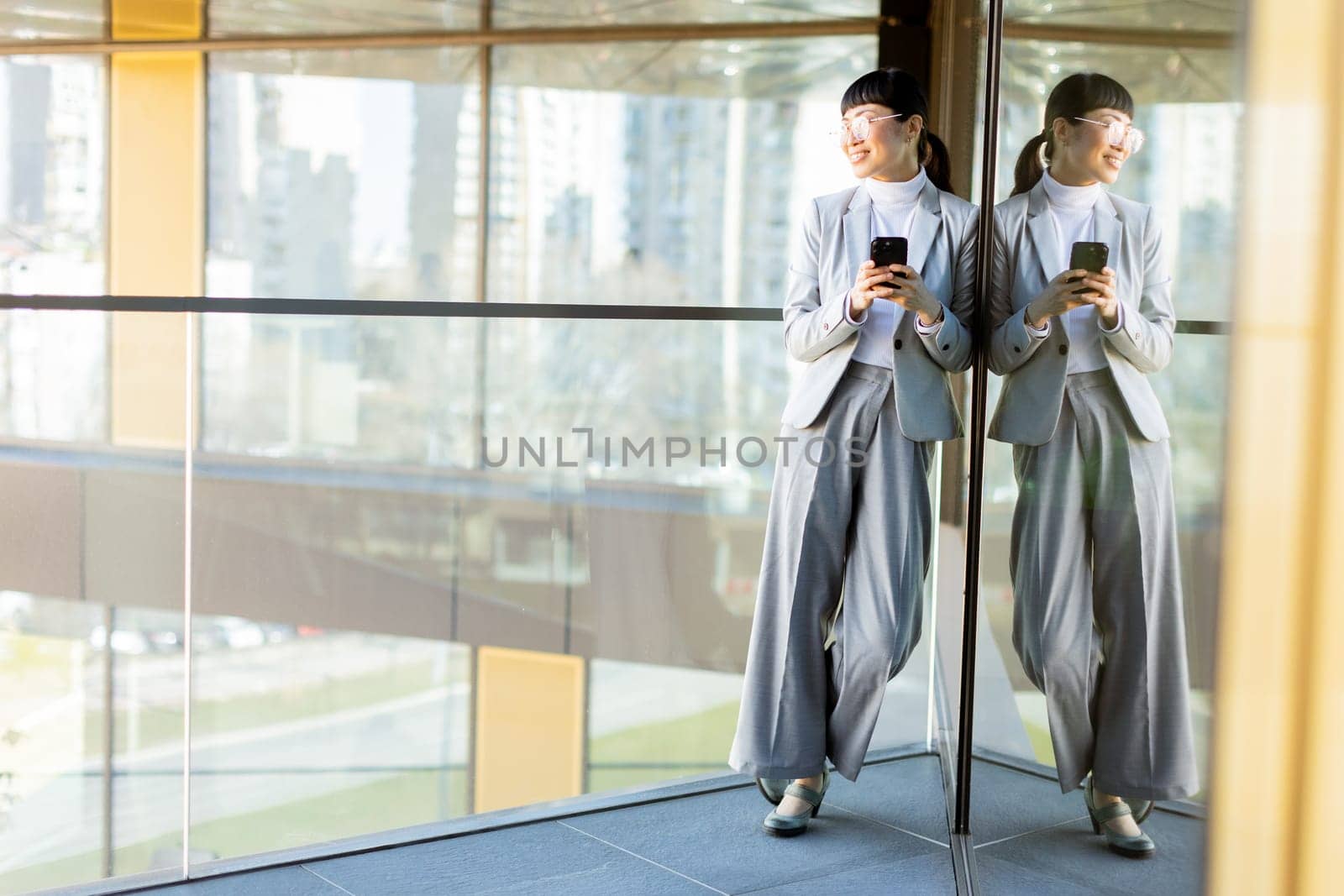 A joyful professional Asian woman dressed in a stylish gray suit smiles widely while holding her phone. She stands in a modern office with large windows reflecting a vibrant cityscape outside.