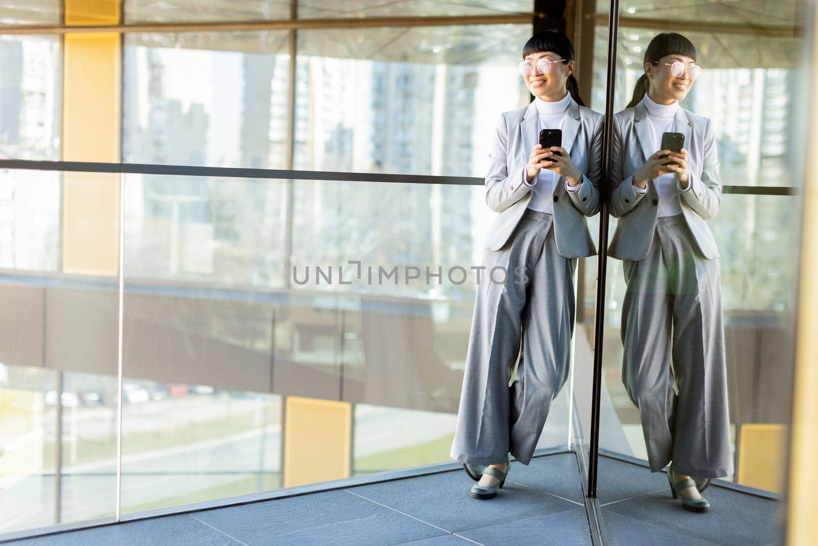 A joyful professional Asian woman dressed in a stylish gray suit smiles widely while holding her phone. She stands in a modern office with large windows reflecting a vibrant cityscape outside.