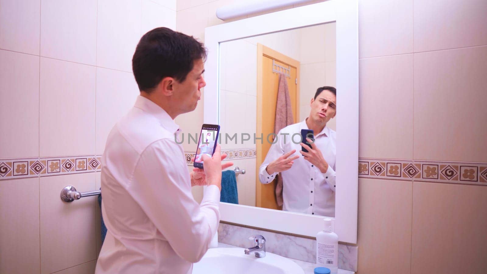Narcissistic businessman taking pictures of himself with smartphone in bathroom mirror, adjusting hair and clothes