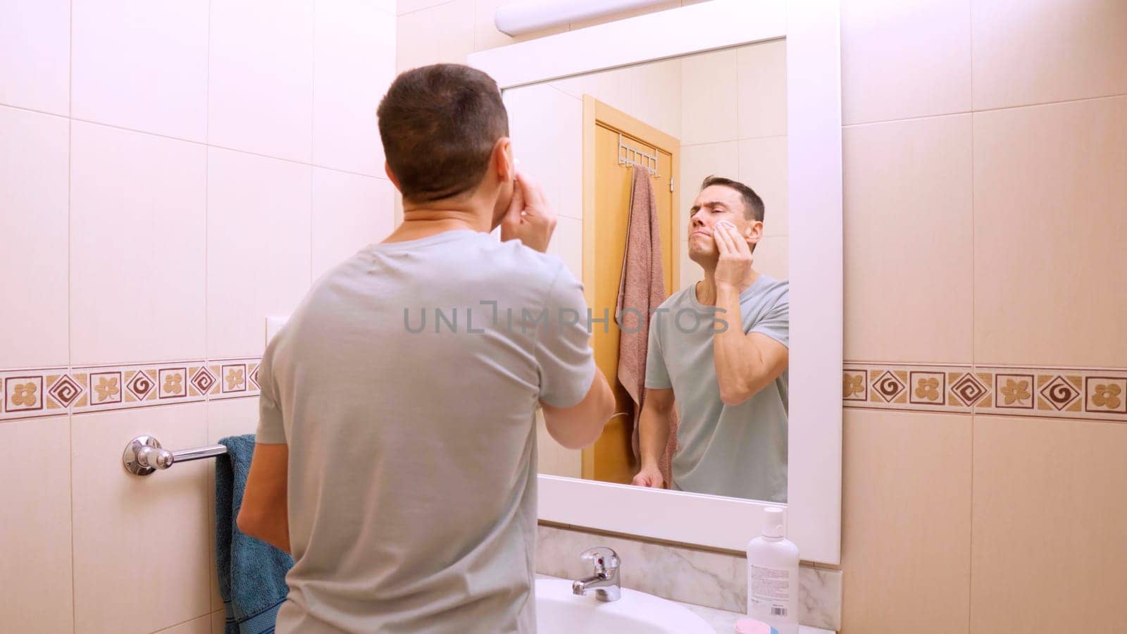 Man performs a detailed facial cleansing routine, using cotton pads and facial products in a tiled bathroom, promoting skincare habits