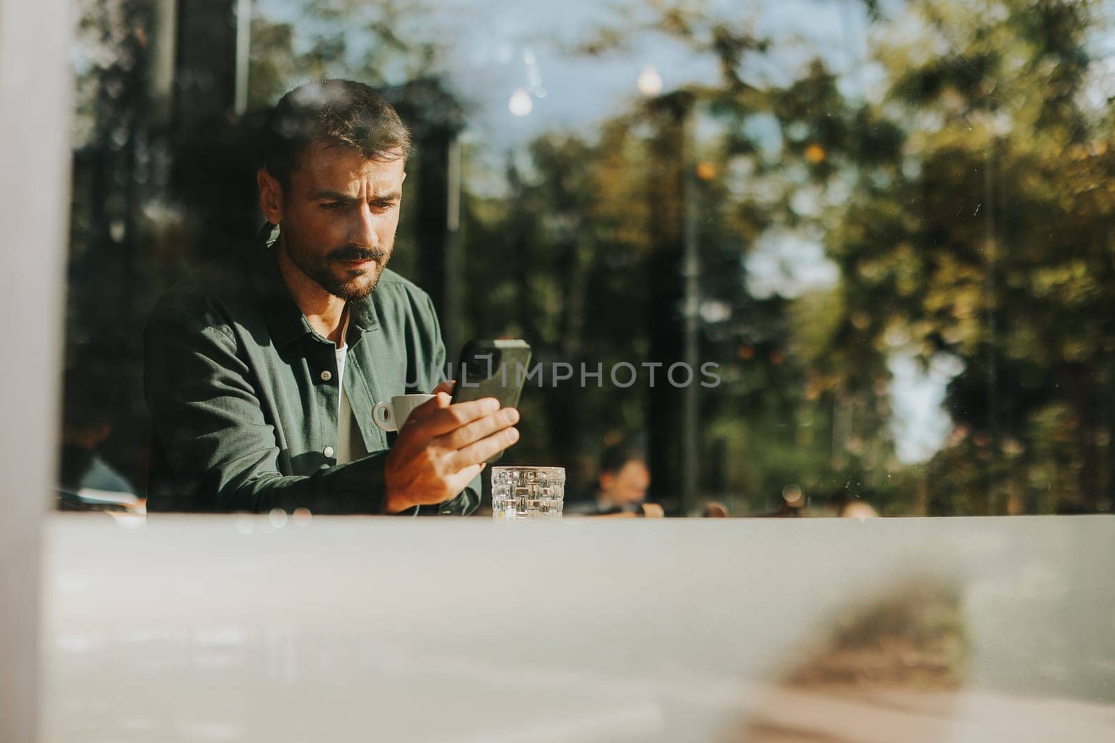In the warm sunlight of a vibrant cafe, a man sits at a table, engrossed in his smartphone, reflecting on modern life amidst the lively atmosphere.