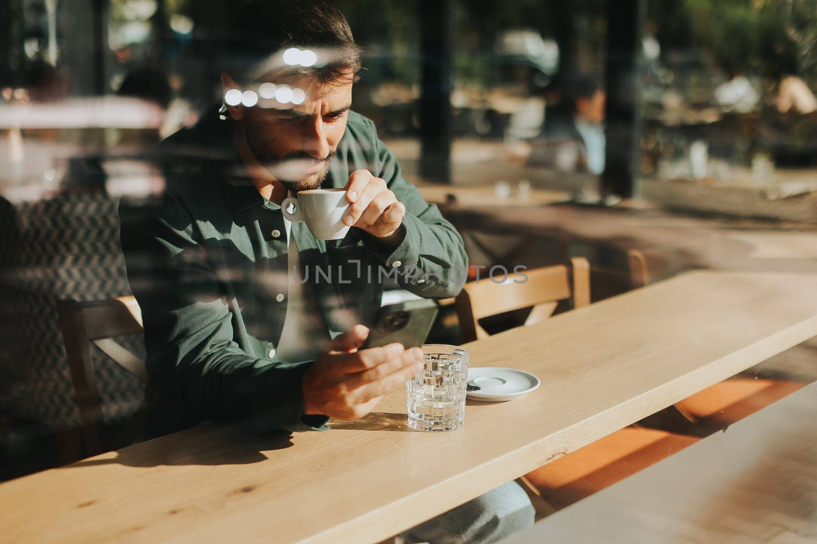 Sitting at a wooden table, a man enjoys his coffee while using mobile phone and gazing out a window, capturing the essence of solitude and reflection in a warm cafe atmosphere
