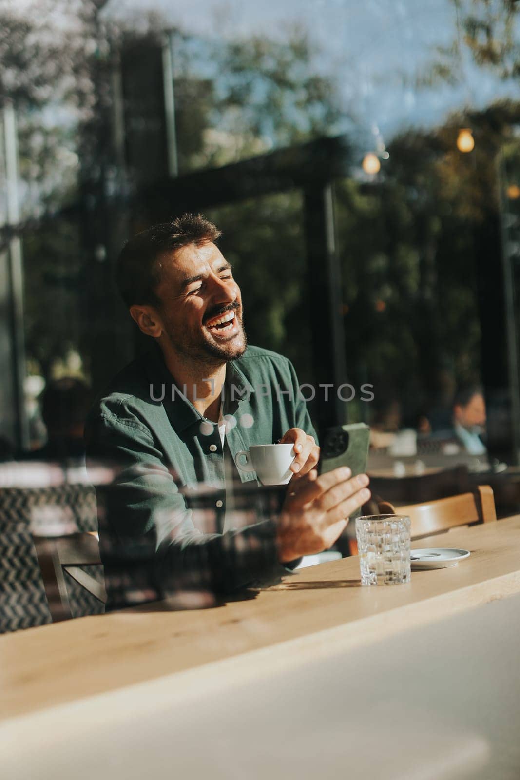 Sitting at a wooden table, a man enjoys his coffee while using mobile phone and gazing out a window, capturing the essence of solitude and reflection in a warm cafe atmosphere