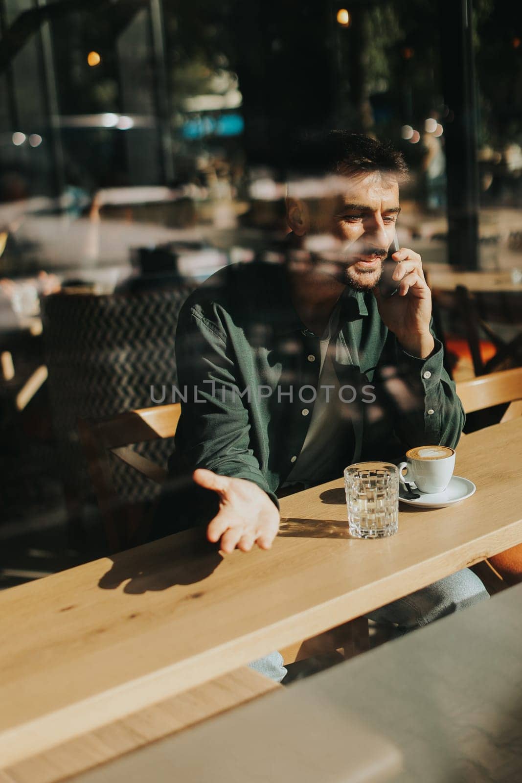 Sitting at a wooden table, a man enjoys his coffee while using mobile phone and gazing out a window, capturing the essence of solitude and reflection in a warm cafe atmosphere