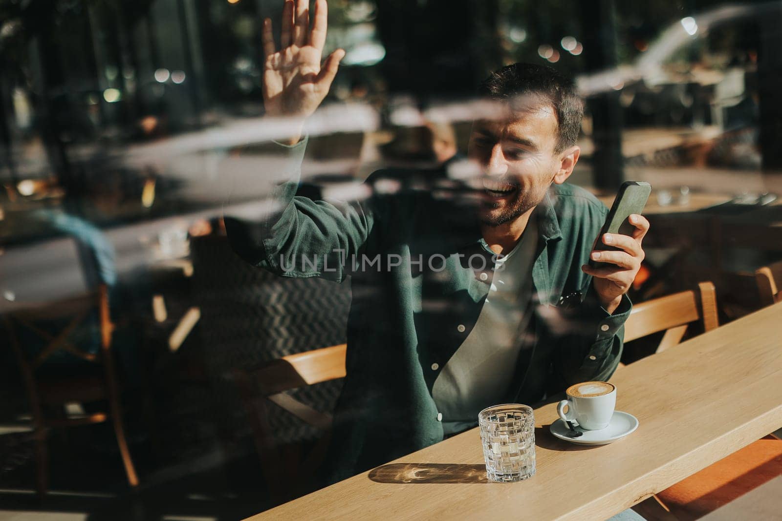 Sitting at a wooden table, a man enjoys his coffee while using mobile phone and gazing out a window, capturing the essence of solitude and reflection in a warm cafe atmosphere