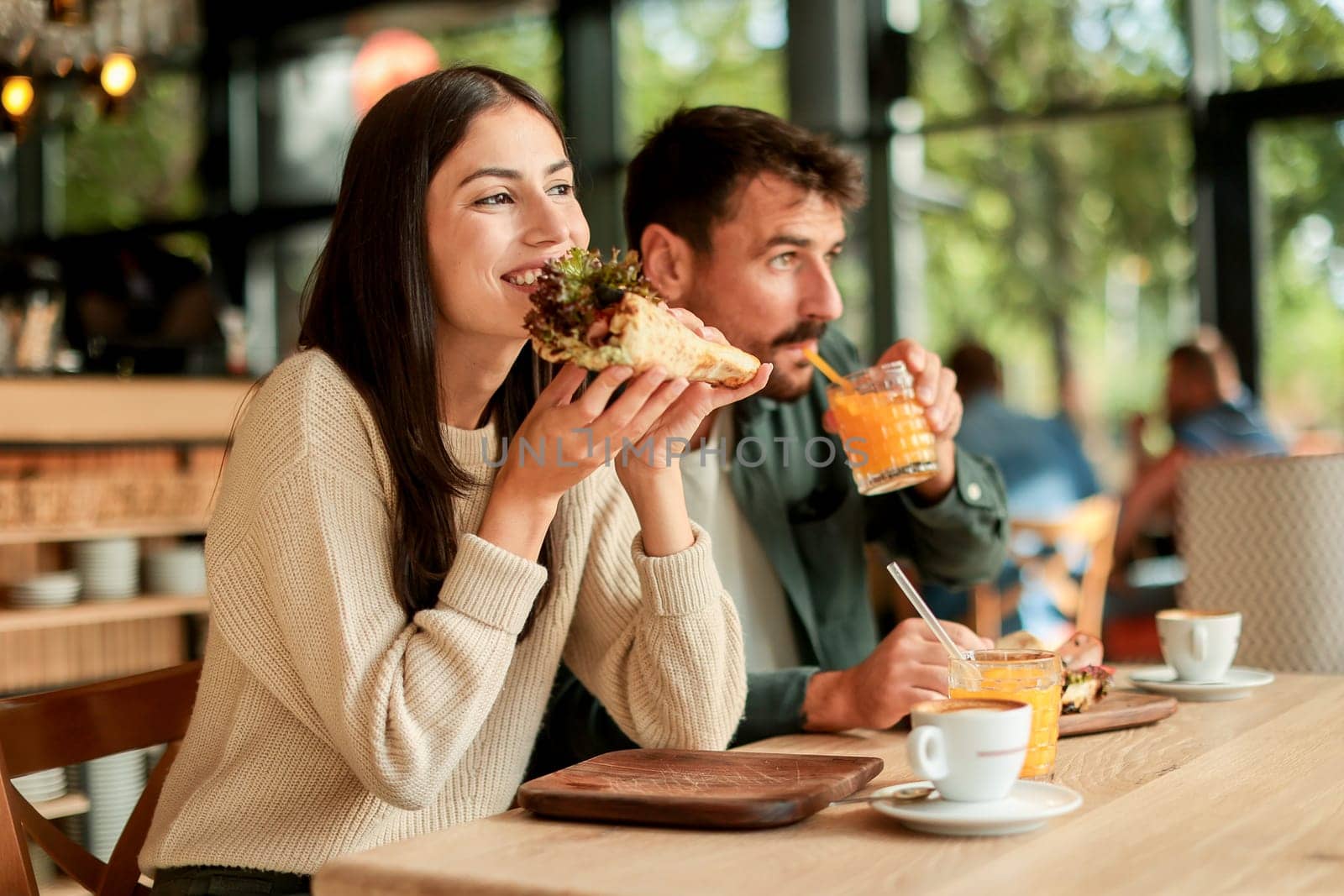 Couple shares laughter and delicious food while sitting at a wooden table in a bright cafe, surrounded by greenery and other patrons enjoying their meals