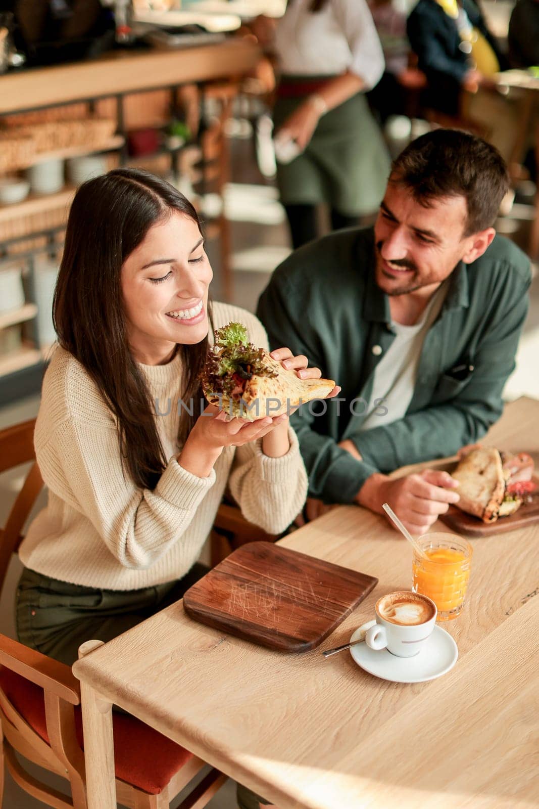 Couple shares laughter and delicious food while sitting at a wooden table in a bright cafe, surrounded by greenery and other patrons enjoying their meals