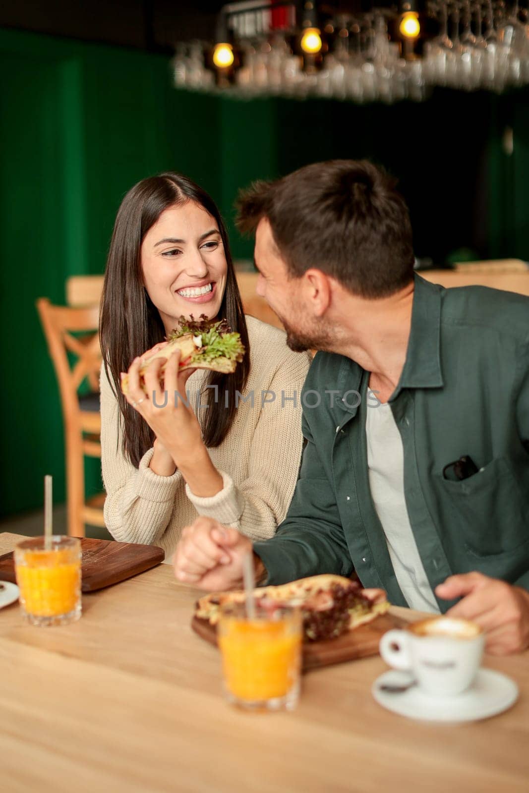 Couple shares laughter and delicious food while sitting at a wooden table in a bright cafe, surrounded by greenery and other patrons enjoying their meals