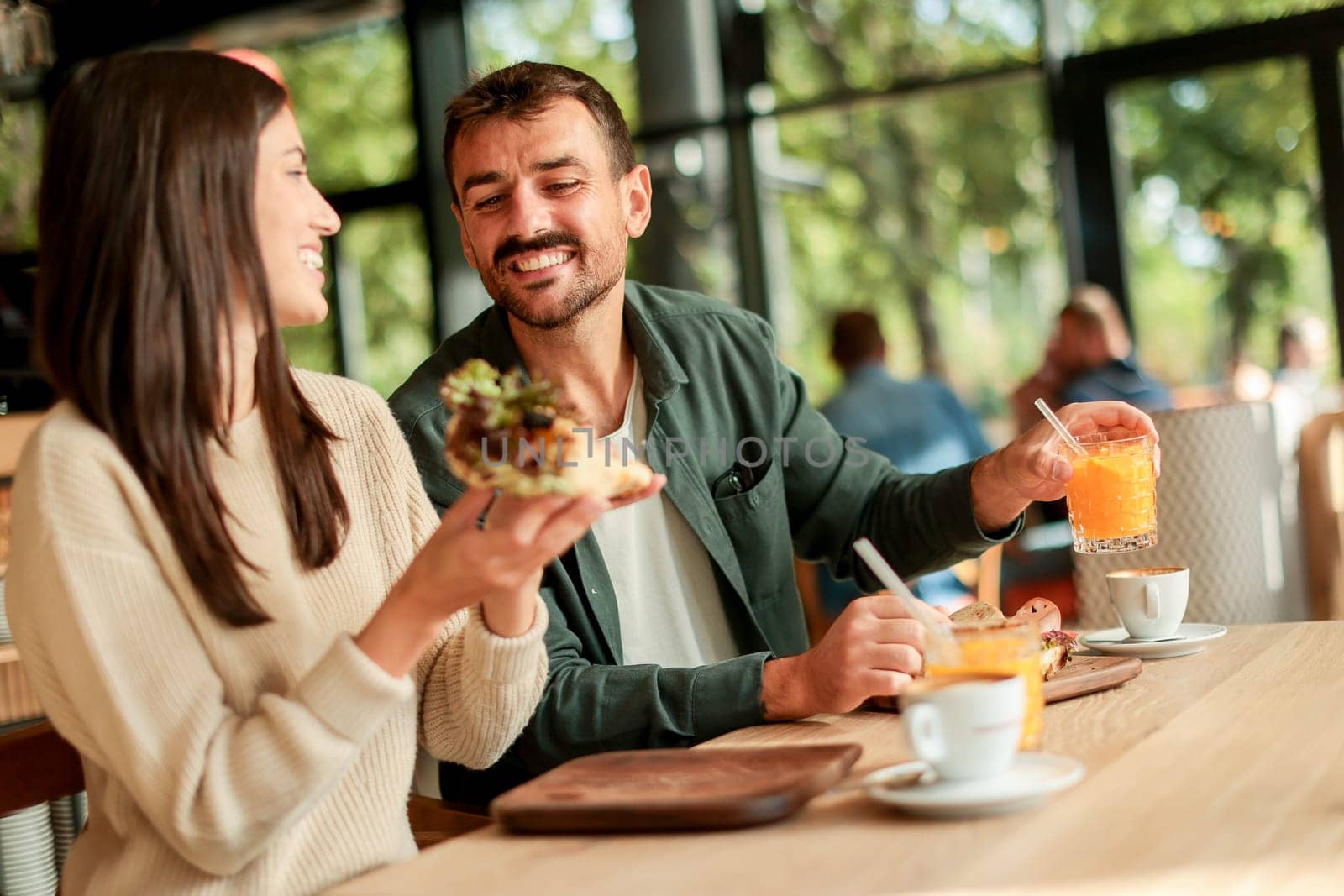 Couple shares laughter and delicious food while sitting at a wooden table in a bright cafe, surrounded by greenery and other patrons enjoying their meals