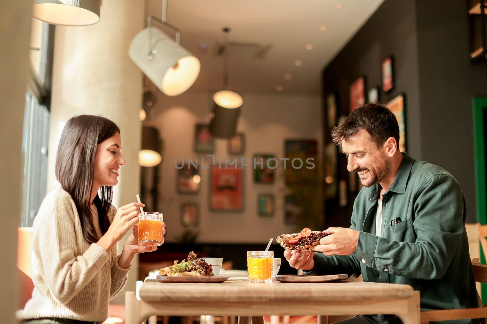 Couple shares laughter and delicious food while sitting at a wooden table in a bright cafe, surrounded by greenery and other patrons enjoying their meals